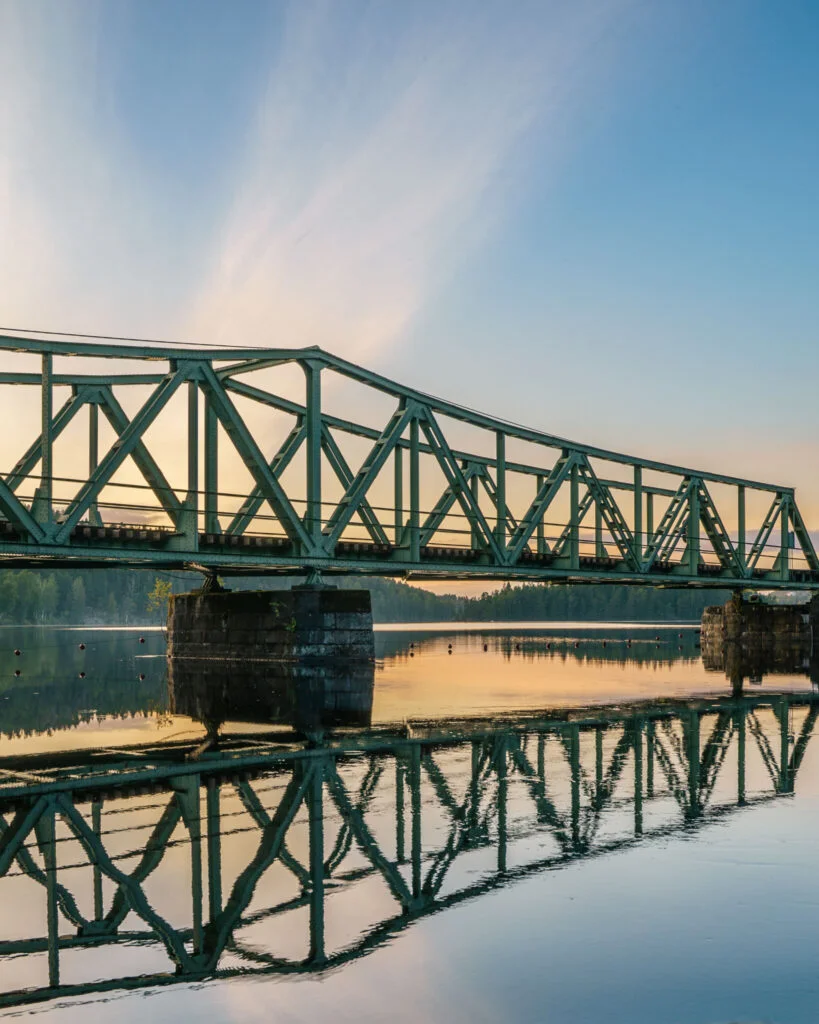 Grön stålbro över Dalslands kanal vid solnedgång, med spegelblank vattenyta som reflekterar brokonstruktionen, nära Bengtsfors