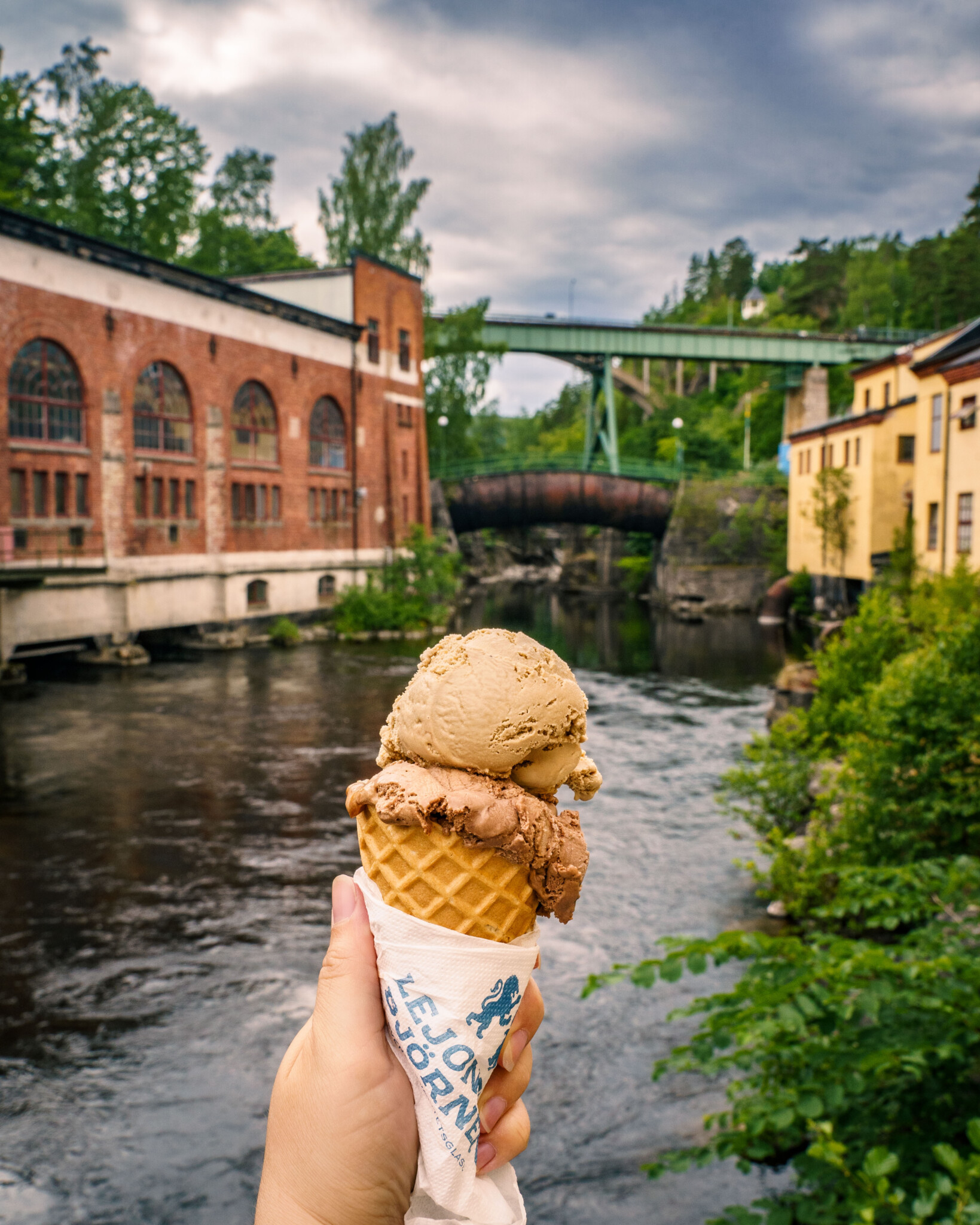 Hand som håller en glasstrut från Lejonet & Björnen med två kulor framför akvedukten i Håverud, med bruket och den gröna bron i bakgrunden
