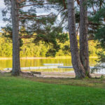 Klöverudsbadet vid Edsleskog – badplats med brygga, sandstrand och gröna gräsytor omgivna av höga tallar vid en stilla sjö.