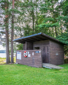 Omklädningshus i brunt trä med skyltar för herrar och damer vid Klöverudsbadet i Edsleskog, med livboj på väggen och sjön skymtande bakom tallarna