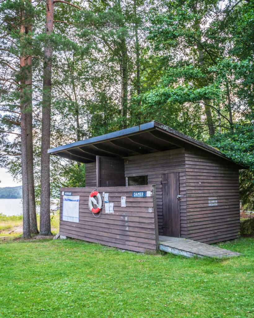 Omklädningshus i brunt trä med skyltar för herrar och damer vid Klöverudsbadet i Edsleskog, med livboj på väggen och sjön skymtande bakom tallarna