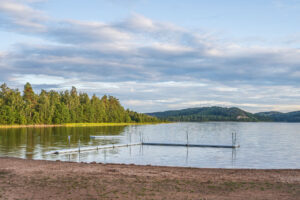 Sandstrand vid Klöverudsbadet i Edsleskog med badbrygga i stilla sjövatten, omgiven av grön skog och kullar i Dalsland.