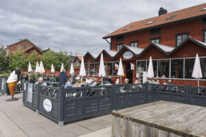 Café Aktersnurran i Nyköpings hamn med uteservering, vita parasoller och en stor glassskylt vid entrén, i en faluröd träbyggnad.