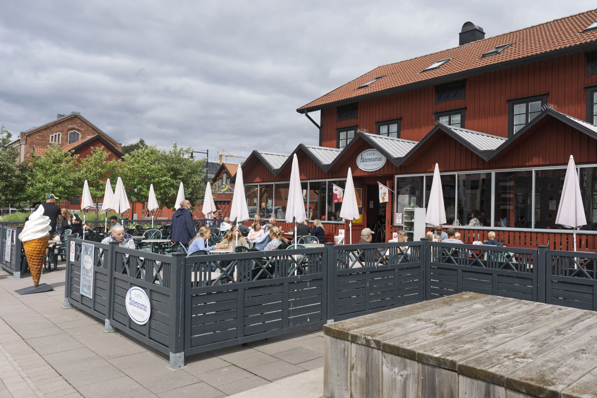 Café Aktersnurran i Nyköpings hamn med uteservering, vita parasoller och en stor glassskylt vid entrén, i en faluröd träbyggnad.