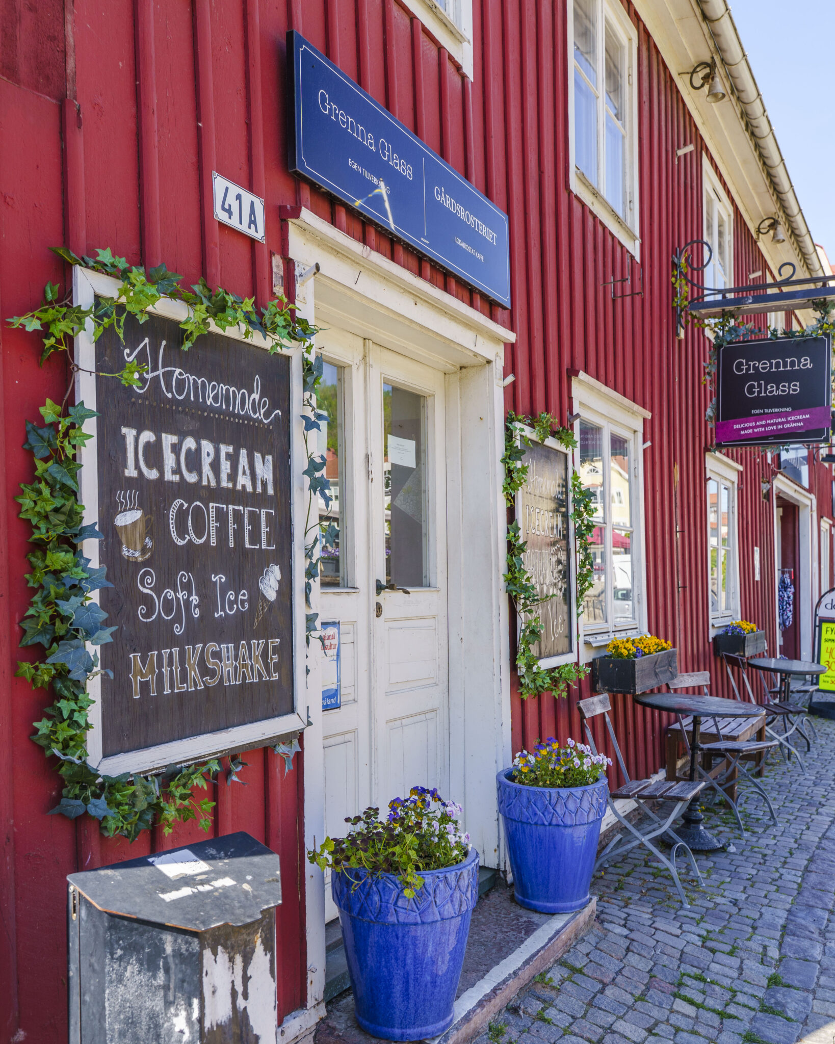Röd träfasad med skylt för Grenna Glass i Gränna, med krittavla som erbjuder hemgjord glass, kaffe och milkshake, blå blomkrukor vid entrén.