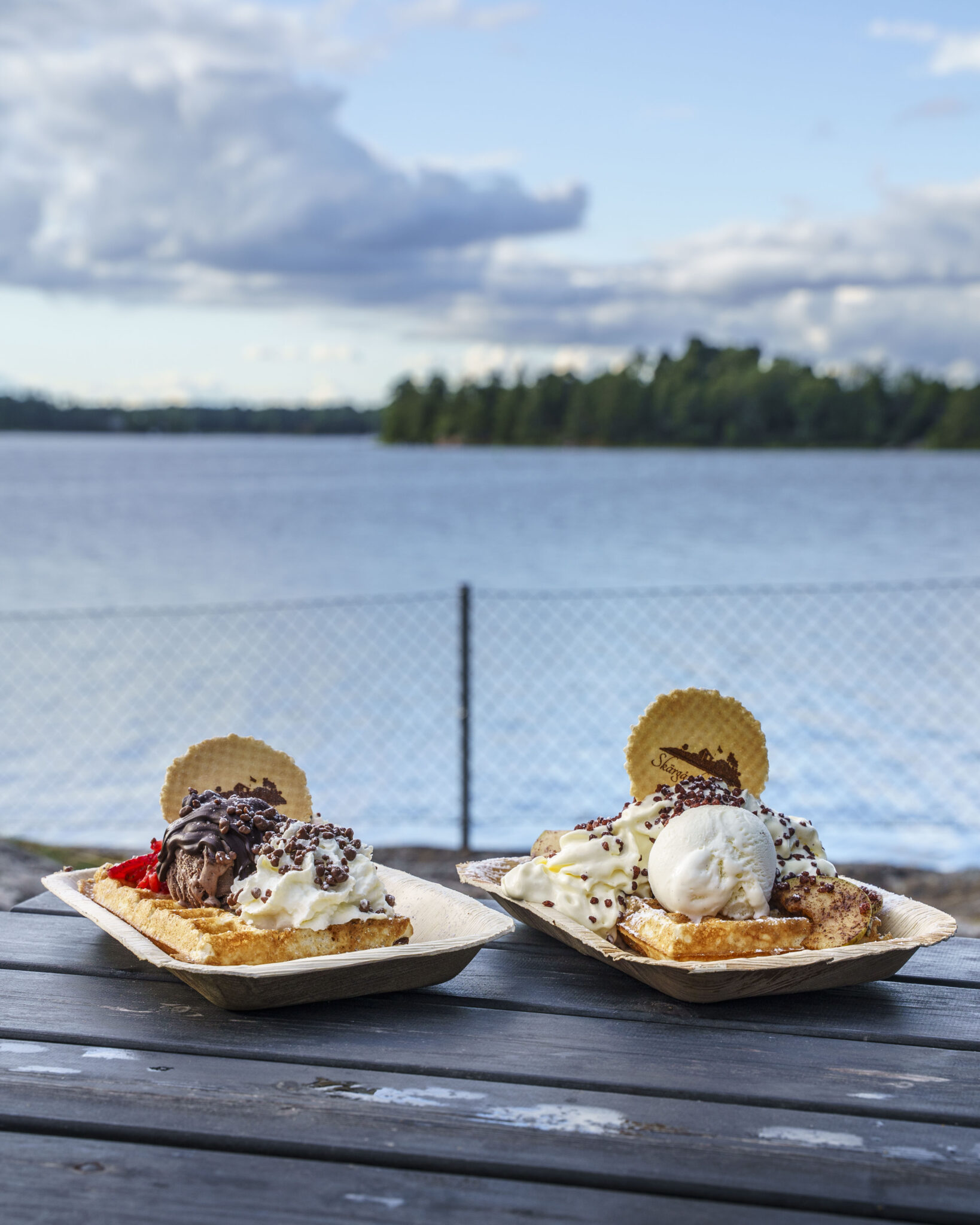 Två dessertvåfflor med glass, grädde och strössel på ett träbord vid vattnet hos Skärgårdsvåfflan i Oxelösund, med skärgårdsvy i bakgrunden.