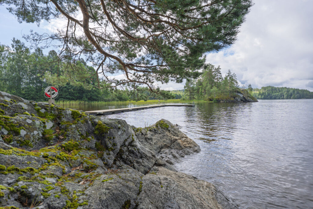 Badplats vid Ivägssjön nära Steneby med mossbeklädda klippor, en flytbrygga och en livboj under en tall i Dalsland.