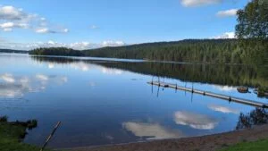 Stilla sjö med spegelblanka reflektioner vid Bengtsbrohöljens badplats, med badbrygga, sandstrand och barrskogsklädd åsrygg i bakgrunden