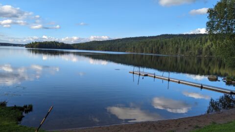 Stilla sjö med spegelblanka reflektioner vid Bengtsbrohöljens badplats, med badbrygga, sandstrand och barrskogsklädd åsrygg i bakgrunden