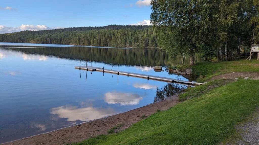 Stilla sjö med badbrygga och sandstrand vid Bengtsbrohöljen, omgiven av gröna gräsytor och barrskog i Dalsland.