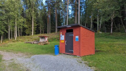 Röd toalettbyggnad med rastplats, grillplats och sittbänkar vid Bengtsbrohöljen badplats, omgiven av barrskog i Dalsland
