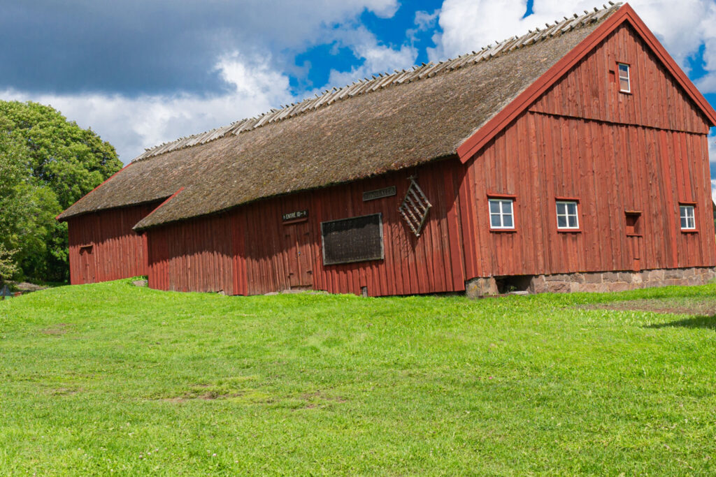 Stor rödfärgad lada med halmtak på Vinnsäters Dalslandsgård, omgiven av grön gräsmatta och lummiga träd i sommarväder.