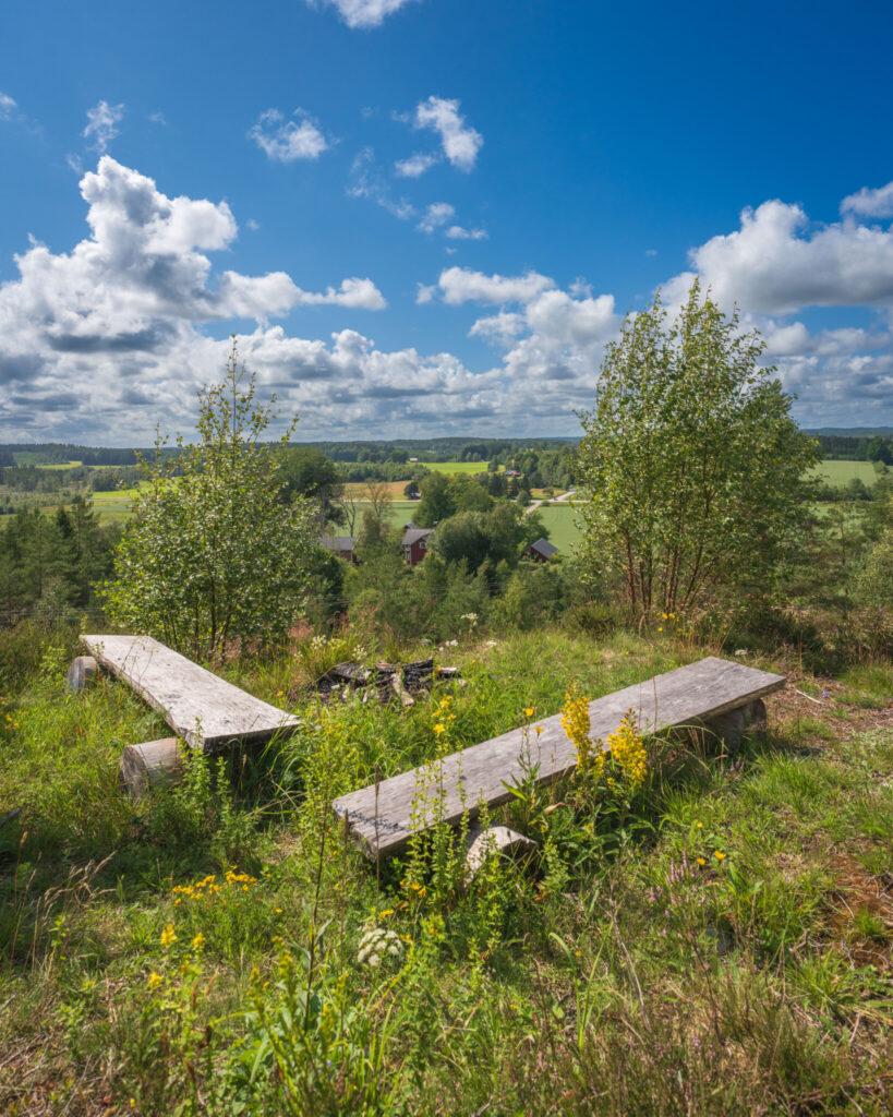 Rustika träbänkar vid eldplats på Tingskullen med vid utsikt över Dalslands gröna jordbrukslandskap och röda stugor en sommardag.