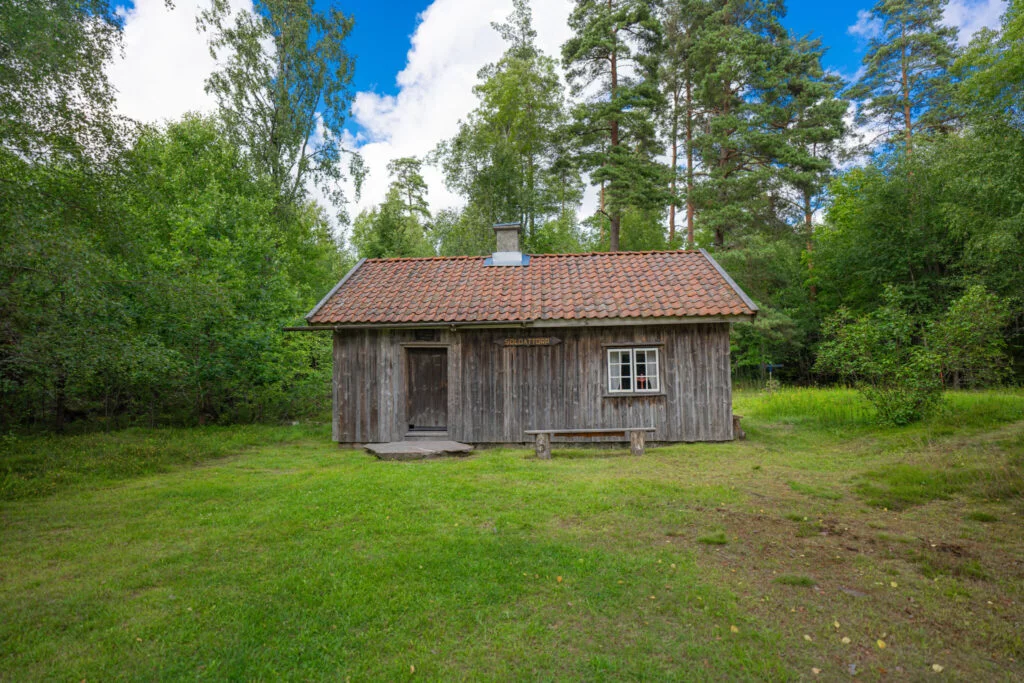 Soldattorpet Bräckan i Järbo, en grå timrad stuga med tegeltak omgiven av grön gräsmatta och lummig skog i Dalsland.
