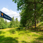 Skylt för Buterudleden (orange led) vid en grönskande stig genom sommargrön skog och äng i Buteruds naturreservat, Dalsland