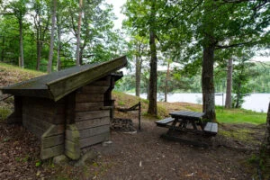 Vindskydd med vedförråd och sittbänk vid sjön i Buteruds naturreservat, omgivet av lövträd och barrskog
