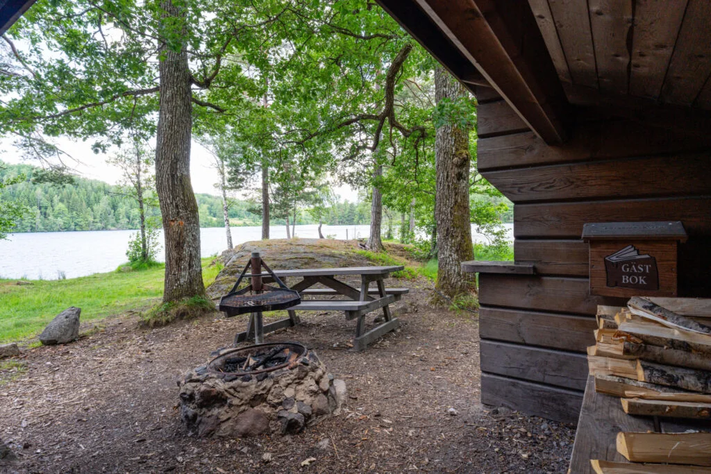 Rastplats vid sjön i Buteruds naturreservat med eldstad, grillgaller, sittbänk, vedtrave och gästbok under tak bland lövträd.