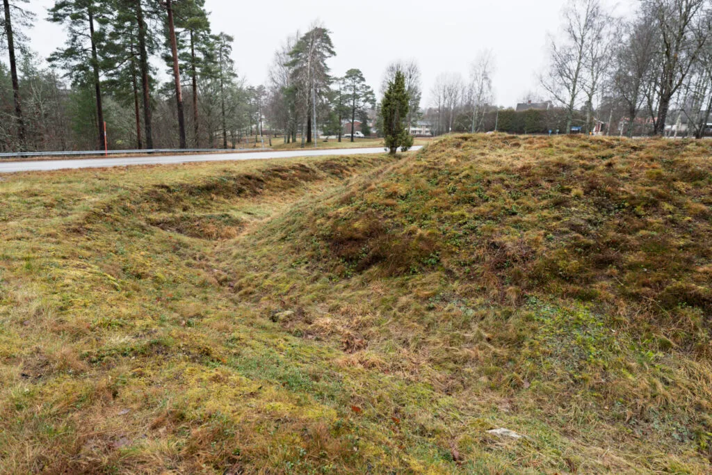 Gräsbevuxna jordvallar och vallgravar vid Redutten i Dals-Ed, en försvarsruin från Napoleonkrigen, med synliga konturer av den forna skansen intill en väg.