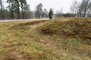 Gräsbevuxna jordvallar och vallgravar vid Redutten i Dals-Ed, en försvarsruin från Napoleonkrigen, med synliga konturer av den forna skansen intill en väg.