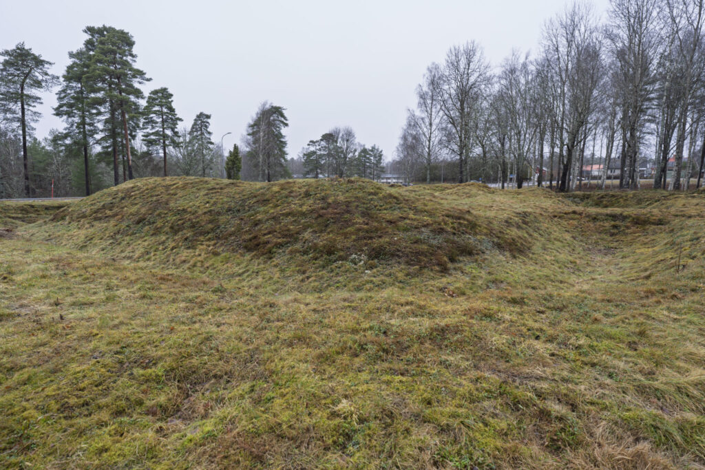 Gräsbevuxna jordvallar vid Redutten i Dals-Ed, en försvarsruin från Napoleonkrigen med synliga konturer av skansens murar, omgiven av träd en grå dag.