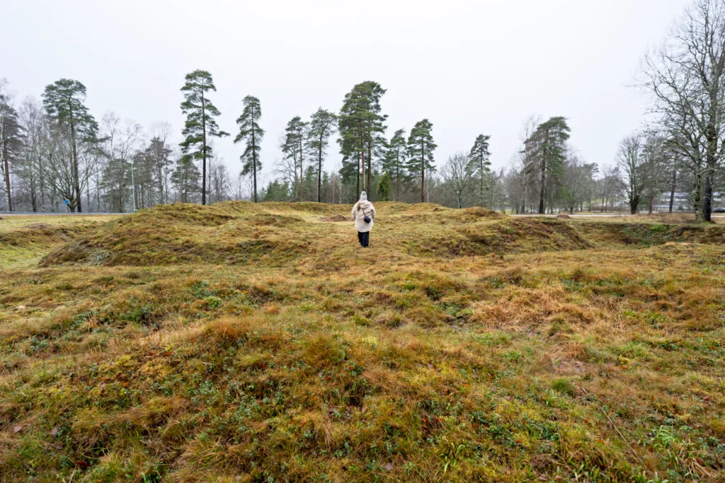 Person går bland gräsbevuxna jordvallar vid Redutten i Dals-Ed, en försvarsruin från Napoleonkrigen med synliga murspår en grå dag.