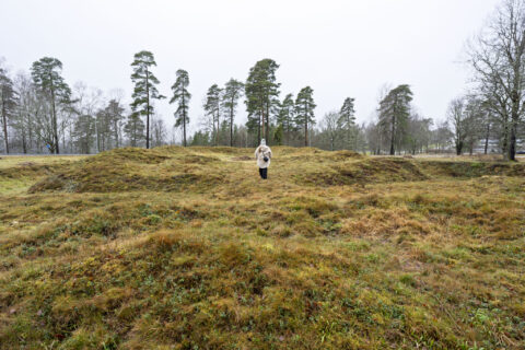 Person går bland gräsbevuxna jordvallar vid Redutten i Dals-Ed, en försvarsruin från Napoleonkrigen med synliga murspår en grå dag.