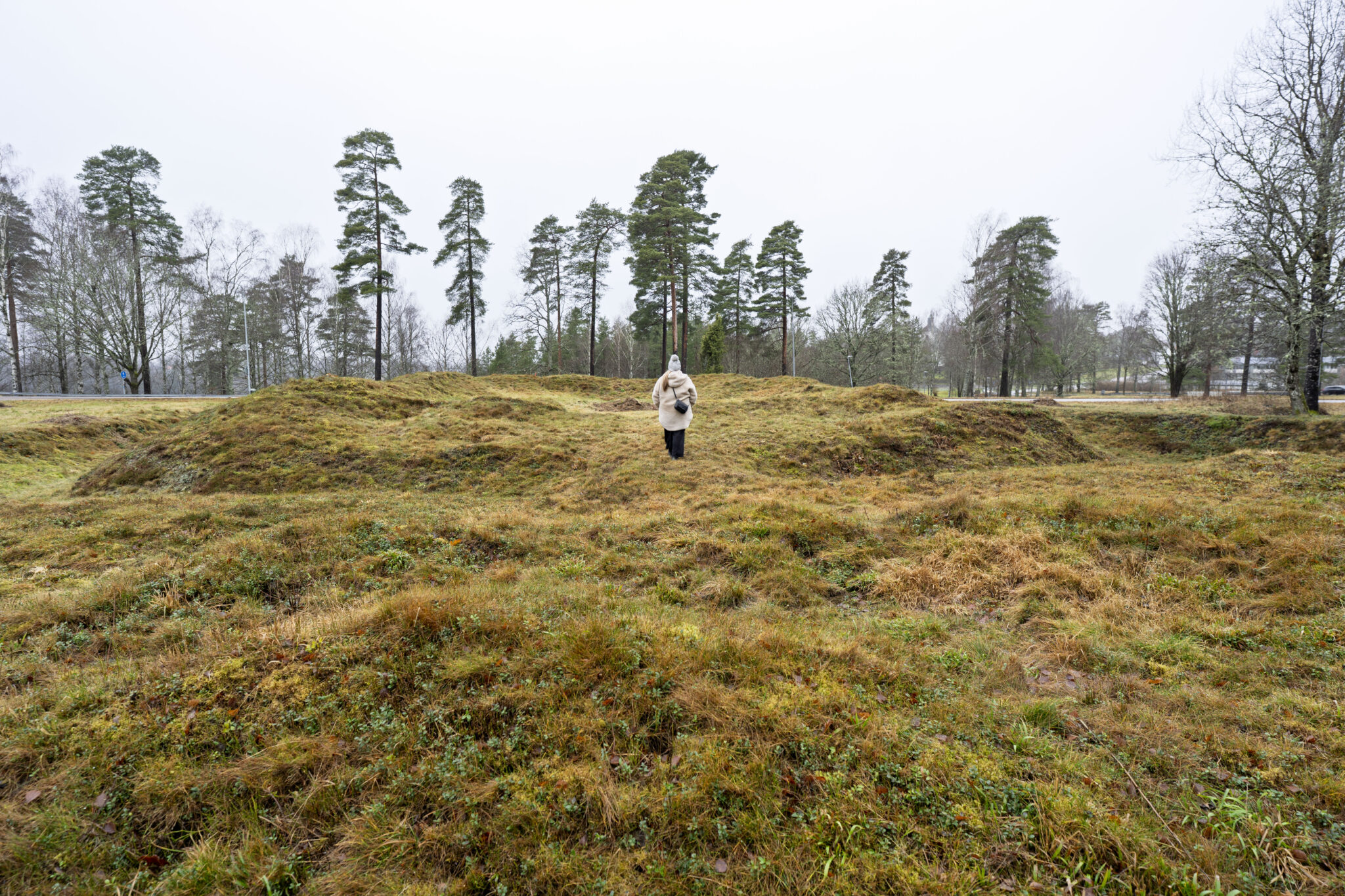 Person går bland gräsbevuxna jordvallar vid Redutten i Dals-Ed, en försvarsruin från Napoleonkrigen med synliga murspår en grå dag.