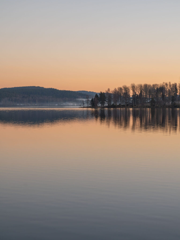 Stilla vintergryning över Laxsjön i Dalsland med spegelblank vattenyta, trädsiluetter och orange himmel.