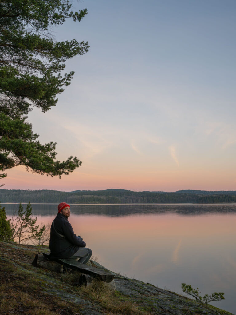 Man med röd mössa sitter på en bänk vid Laxsjöns strand i solnedgång, omgiven av tallar i Ösans naturreservat, Dalsland.