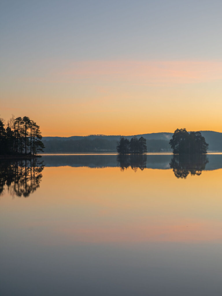 Spegelblank solnedgång över Laxsjön i Dalsland med siluetter av tallbevuxna öar reflekterade i det stilla vattnet.