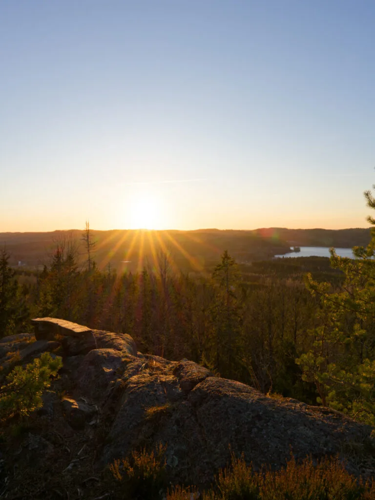 Solnedgång över skogslandskapet i Åmålsbergen med klippor i förgrunden och en glittrande sjö i fjärran.