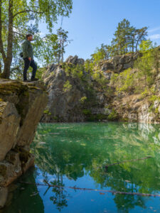 Man står på en klippavsats och blickar ut över Bläsens blå lagun, ett vattenfyllt gruvhål med smaragdgrönt vatten omgivet av berg och skog i Dalsland.