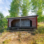 Faluröd vindskydd vid Trehörnabadet längs Östgötaleden, omgivet av björkar och grönska i sommarlandskap.
