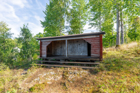 Faluröd vindskydd vid Trehörnabadet längs Östgötaleden, omgivet av björkar och grönska i sommarlandskap.