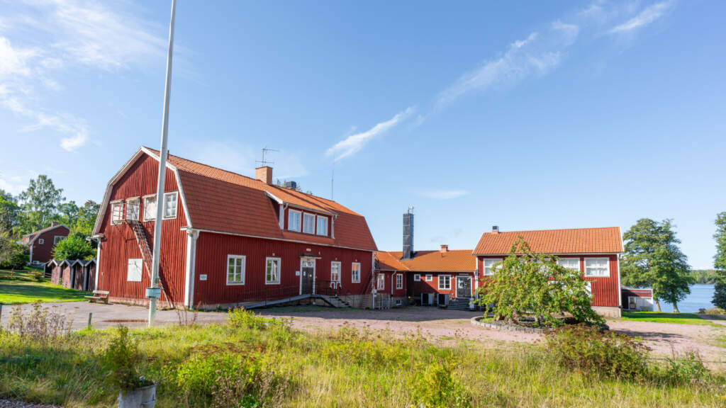 Röda träbyggnader vid Trehörna Wärdshus i Östergötland med flaggstång och sjön synlig i bakgrunden en solig sommardag