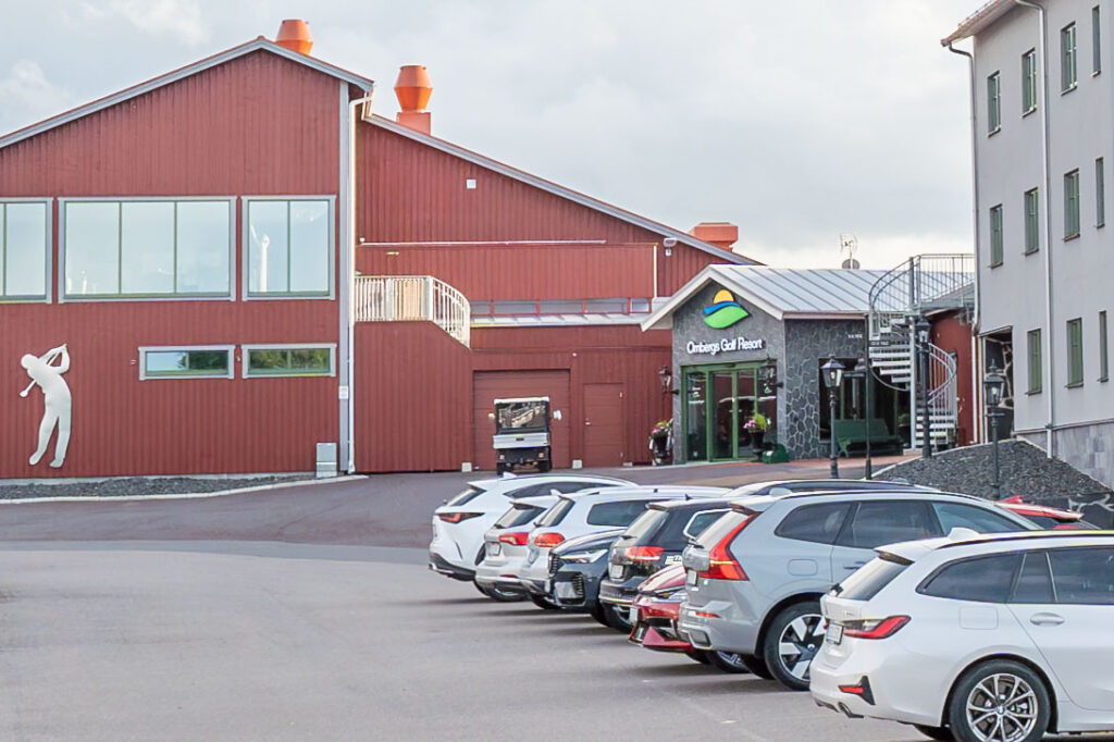 Ombergs Golf Resorts entré med röd träbyggnad, golfarsilhuett på fasaden och parkerade bilar framför under en mulen himmel.