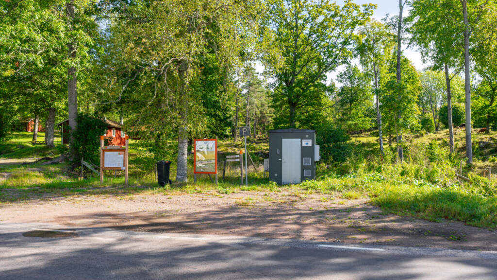 Parkering vid Östgötaleden i Trehörna med informationstavlor, toalett och soptunna omgiven av lummig lövskog sommartid.