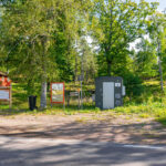 Parkering vid Östgötaleden i Trehörna med informationstavlor, toalett och soptunna omgiven av lummig lövskog sommartid.