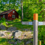 Vägvisare på Östgötaleden med pilar mot Ödeshög och Boxholm, framför en röd stuga omgiven av grön lövskog vid Trehörna