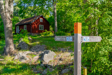 Vägvisare på Östgötaleden med pilar mot Ödeshög och Boxholm, framför en röd stuga omgiven av grön lövskog vid Trehörna