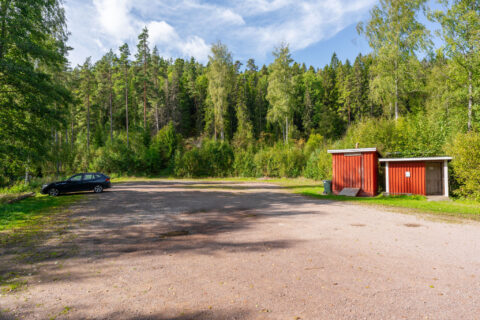Grusad parkering vid Klintens badplats med en parkerad bil, röd toalettbyggnad och tät barrskog i bakgrunden, Ödeshög