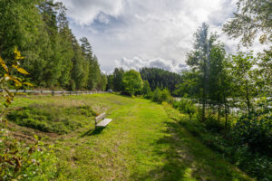 Grön gräsyta vid Klintens badplats med en träbänk, omgiven av lummig skog och med Vagnsjön skymtande till höger.