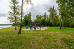Lekplats med klätterställning och rutschkana på grönytan vid Visjö badplats, omgiven av björkar och grönska nära sjön.