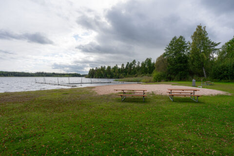 Visjö badplats med sandstrand, picknickbänkar på gräsmatta och badbryggor i sjön, omgiven av skog en mulen dag.