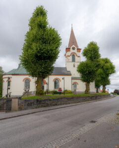 Ödeshögs kyrka med vit putsad fasad, spetsigt klocktorn och rundbågade fönster, omgiven av gröna lövträd längs en gata en mulen dag.