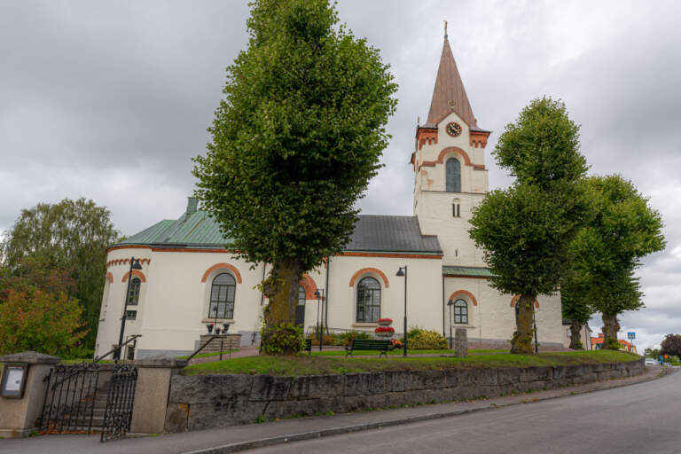 Ödeshögs kyrka med vit fasad, rundbågade fönster och spetsigt torn, omgiven av gröna lövträd en mulen dag