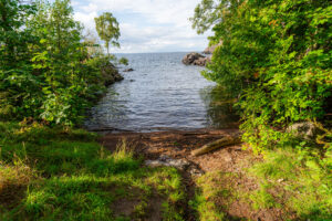 Liten naturlig vik vid Öninge hamn längs Vätterns kust, omgiven av frodig grönska och klippor, med stig ner till vattnet.