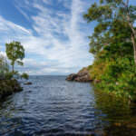 Klippor och grönskande lövträd ramar in en stilla vik vid Öninge hamn längs Vätterns kust nära Ödeshög, under en blå himmel med slöjmoln.