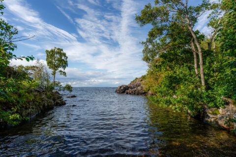 Klippor och grönskande lövträd ramar in en stilla vik vid Öninge hamn längs Vätterns kust nära Ödeshög, under en blå himmel med slöjmoln.