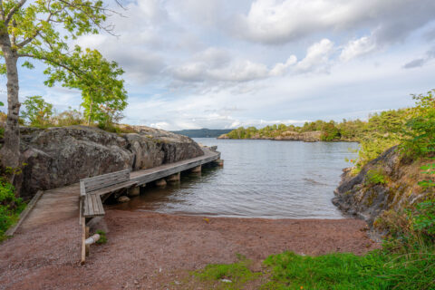 Skyddad badvik med träbrygga vid klipporna i Stora Lunds naturreservat, Vättern i bakgrunden under molnig himmel.